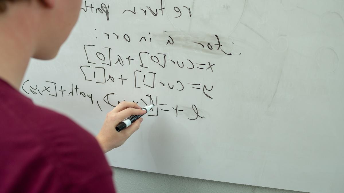 A student writes code and math on a whiteboard. 